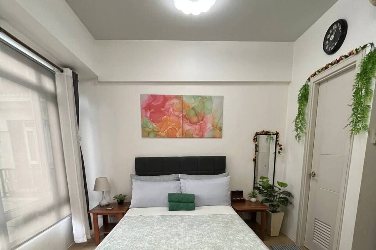 Un dormitorio con cama, un banco, una mesita de noche, una lámpara, un reloj y una ventana con cortinas.