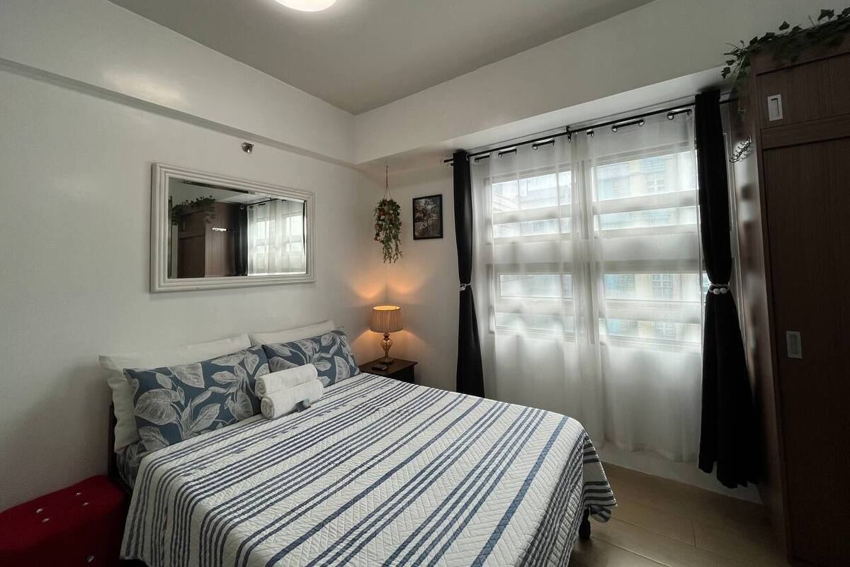 Un dormitorio con cama, lámpara de noche, espejo y ventana con cortinas.