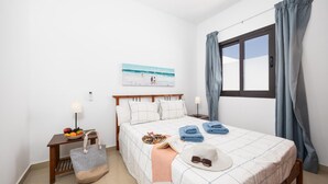 A neatly arranged bedroom with a bed, bedside tables, a fruit bowl, and a beach-themed painting.