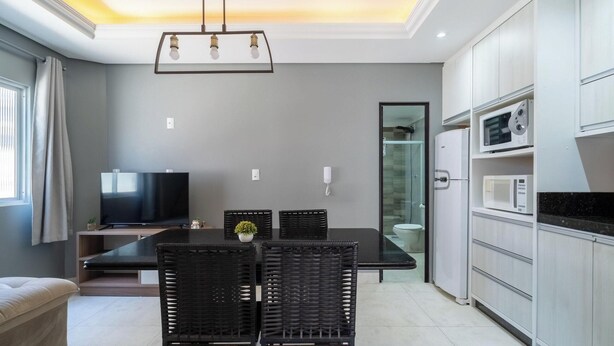 A modern kitchen with a dining area, a flat-screen TV, and a glass shower enclosure. - Apts Near the Beach w/ Full Amenities - RNC (Florianópolis)