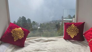 Una cama con dos almohadas rojas con diseños dorados, una colcha blanca y vistas a árboles y edificios a través de una ventana.