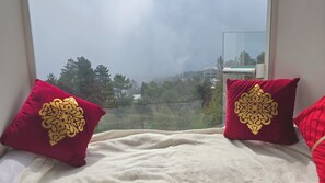 Una cama con dos almohadas rojas con diseños dorados, una colcha blanca y vistas a árboles y edificios a través de una ventana.