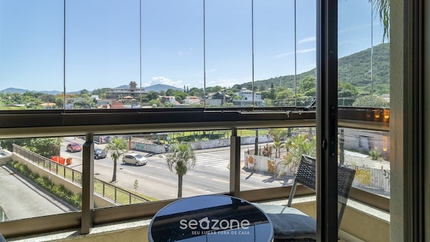 Vista da janela com moldura de metal e vidro, mostrando uma rua com carros, palmeiras e edifícios.