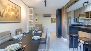 Uma zona de refeições moderna com uma mesa comprida posta para seis pessoas, uma cozinha com armários de madeira e uma área de bar com banquetas.