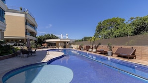 Piscina com espreguiçadeiras e área de estar coberta, cercada por um edifício com telhado plano e um carro estacionado por perto.
