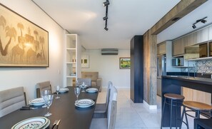 Un espace repas moderne avec une longue table dressée pour six personnes, une cuisine dotée de meubles en bois et un coin bar avec des tabourets.