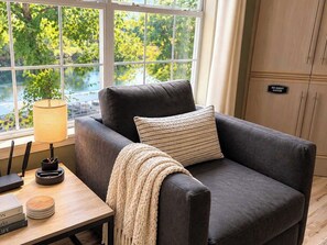 A dark-colored sofa with a knitted throw blanket and a striped pillow, positioned next to a window with a view of trees.