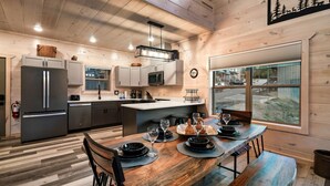 A modern kitchen with a wooden interior, a dining area with a wooden table set for four, and a large window offering a view of the outdoors. - NoBunks! 7Beds•Spacious & Near Parkway/Attractions (Pigeon Forge)