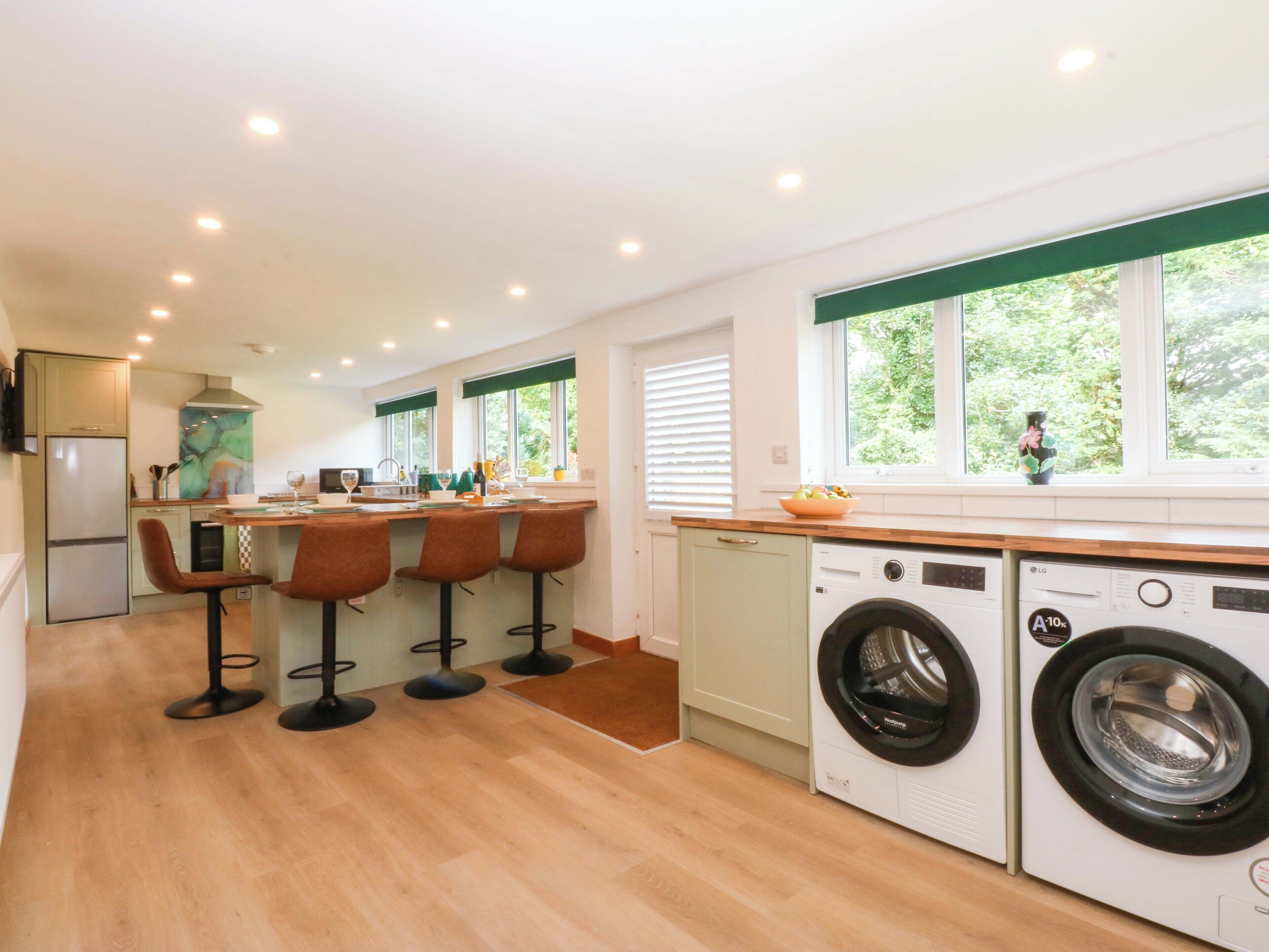 A modern kitchen with a central island, bar stools, a washing machine, and a large window with a view of greenery.