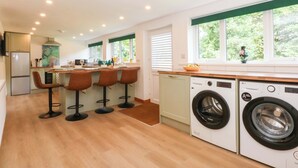 A modern kitchen with a central island, bar stools, a washing machine, and a large window with a view of greenery. - Cromlech Cottage at Cromlech Manor Farm (Tyn-y-Gongl)