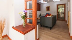 A living room with a grey sofa, a wooden side table with a vase of flowers, and a view of a dining area through an open door. - Cromlech Cottage at Cromlech Manor Farm (Tyn-y-Gongl)