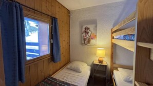 Et lille, hyggeligt værelse med trævægge, en enkeltseng, et vindue med gardiner, en sengebordlampe og en køjeseng.