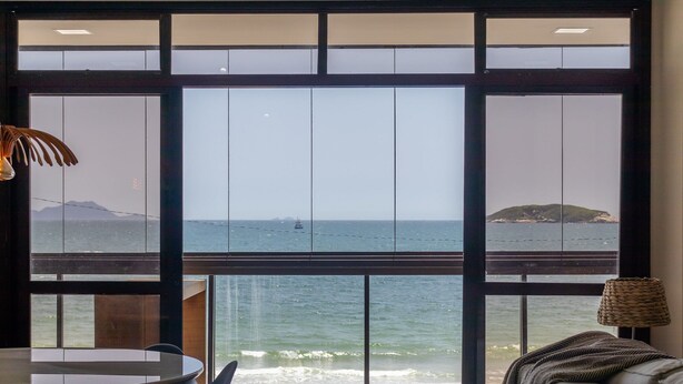 A room with a large window overlooking a beach, a round table, and a sofa with a lamp. - Beautiful Seaside Apartment With Barbecue Rig0102 (Florianópolis)
