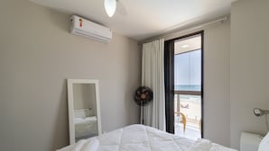 Ein Hotelzimmer mit Bett, großem Spiegel, Klimaanlage, Ventilator, Fenster mit Blick auf den Strand und Nachttisch mit Lampe.