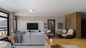 A modern living room with a large flat-screen TV, a sofa, a wicker chair, and a dining table.