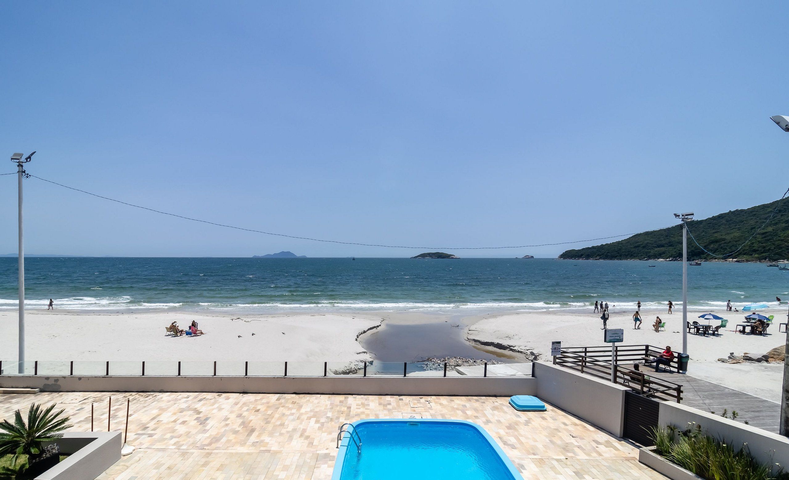 Vista a la playa desde una zona de piscina, con cielo despejado, arena y algunas personas a lo lejos.