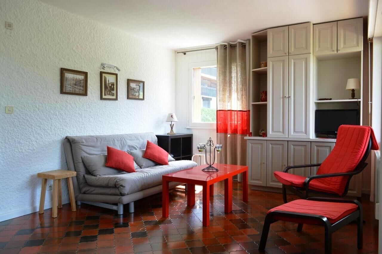 Un salon avec un canapé, une table rouge, une chaise rouge et un téléviseur. On y trouve également des armoires et une fenêtre avec des rideaux.