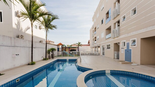 A swimming pool with a curved design, surrounded by a building with balconies and a playground in the background. - Apt 500m From Ingleses Beach, Floripa Rid3106 (Florianópolis)