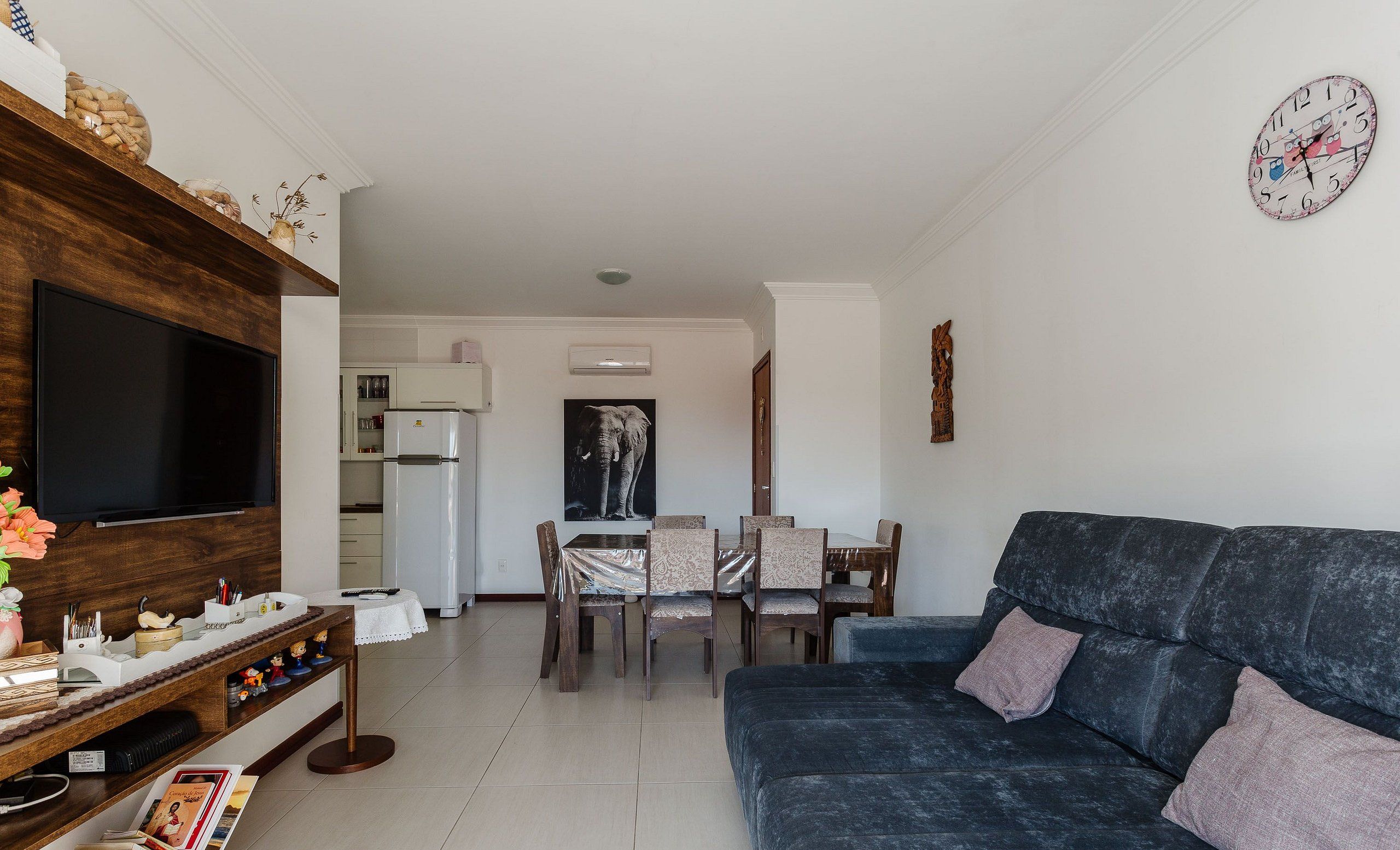 Una sala de estar moderna con un televisor de pantalla plana, un sofá azul oscuro, una zona de comedor con sillas y una mesa, y una cocina con refrigerador y armarios.