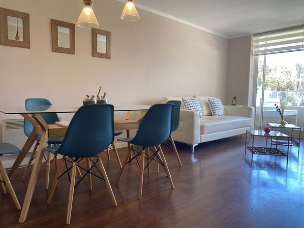Una sala de estar moderna con comedor, que incluye un sofá blanco, sillas azules, una mesa de vidrio y suelo de madera.