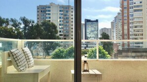 A balcony with a white chair, a small table, and a bottle. The balcony overlooks a cityscape with multiple high-rise buildings and trees.