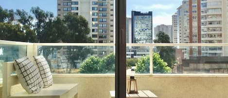 A balcony with a white chair, a small table, and a bottle. The balcony overlooks a cityscape with multiple high-rise buildings and trees.