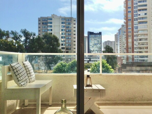 A balcony with a white chair, a small table, and a bottle. The balcony overlooks a cityscape with multiple high-rise buildings and trees.