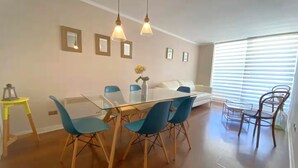 A dining area with a glass table, blue chairs, and pendant lights. There is a sofa and a window with blinds in the background.