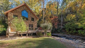 Une cabane en pierre avec une terrasse en bois, entourée d’un feuillage automnal et bordée par une rivière.