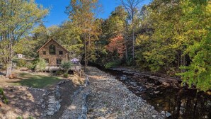 Un chalet rustique en pierre, doté d’une terrasse en bois, entouré d’une zone boisée et bordé par un ruisseau.