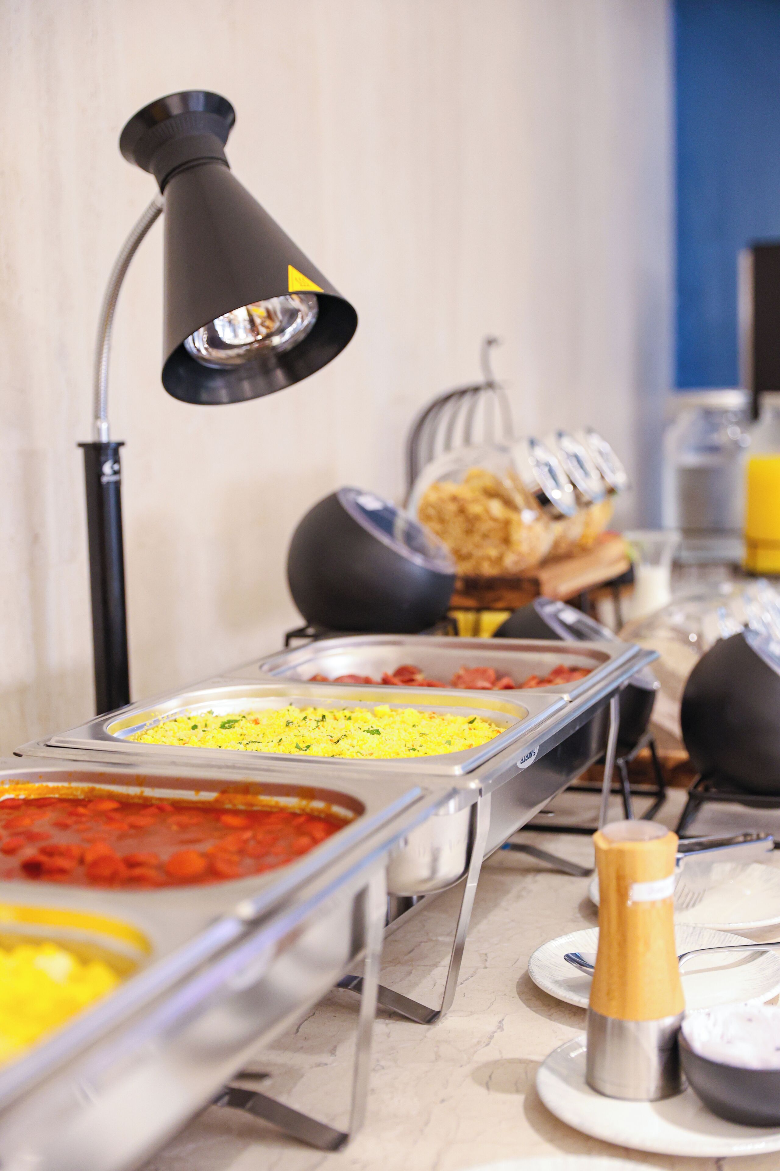 A buffet setup with various food items in trays, a black lamp, and condiments on the counter.