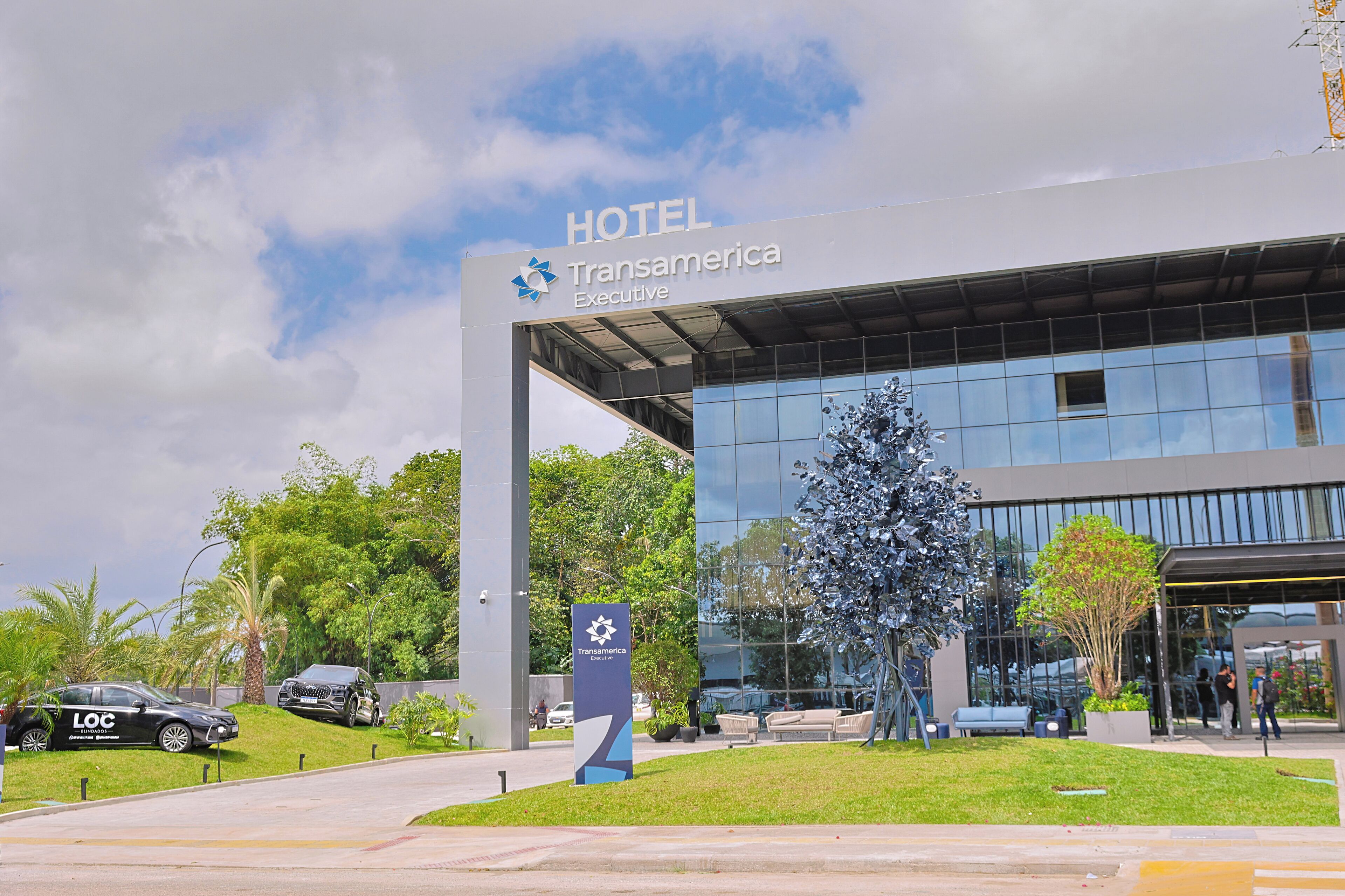 A hotel entrance with a modern glass facade, a sign for Transamerica Executive, and a landscaped area with a tree and benches.