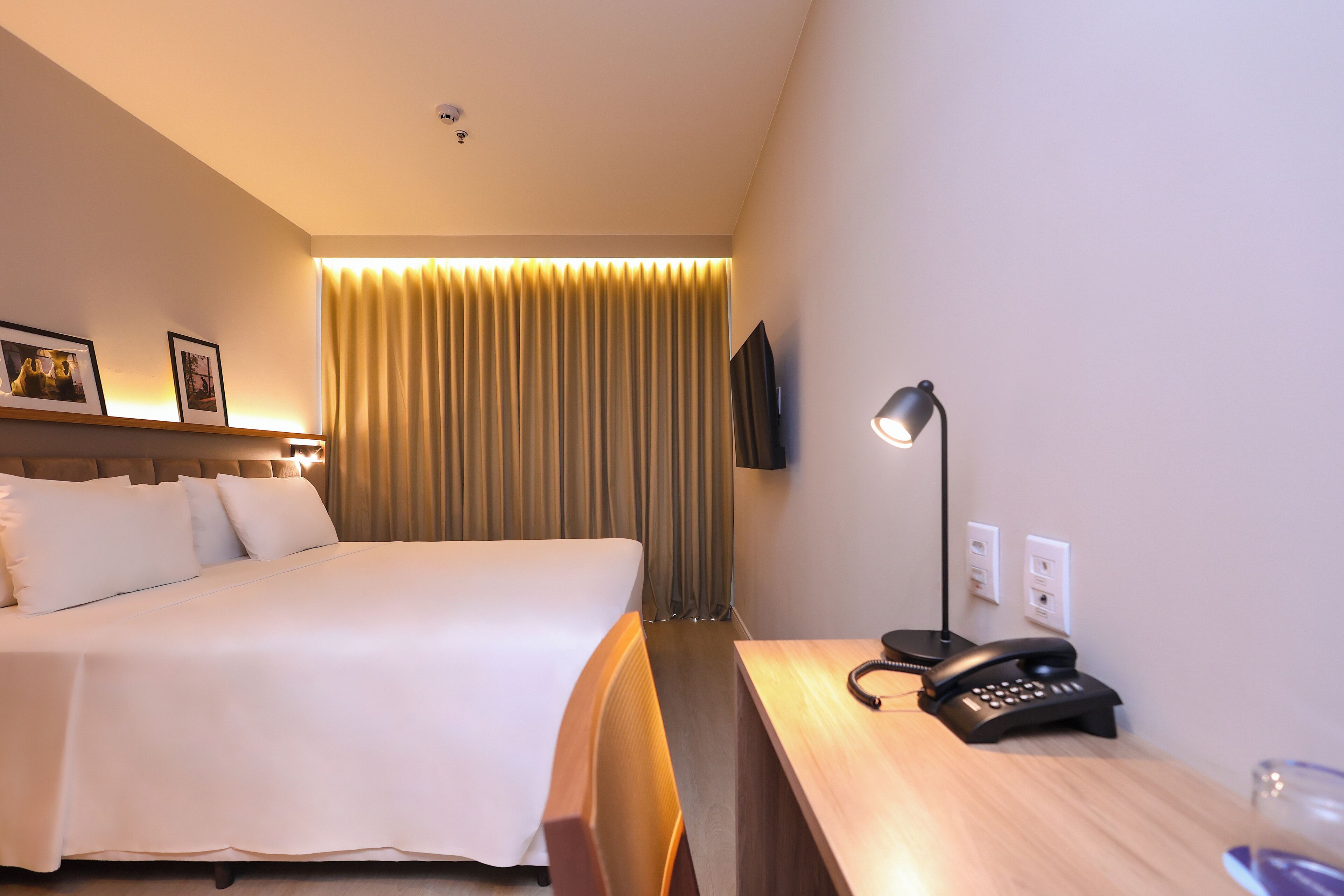Una habitación de hotel con una cama grande, un escritorio con teléfono, una lámpara y un televisor montado en la pared.