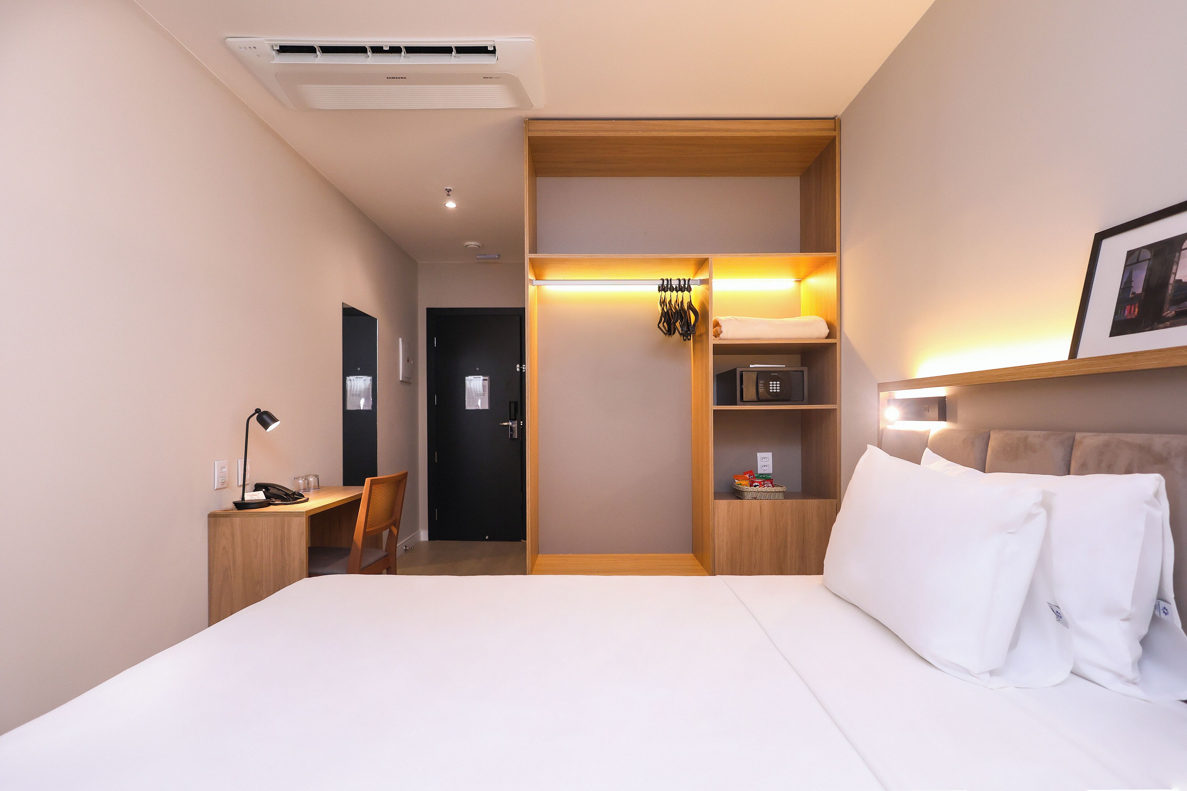 Una habitación de hotel con cama, escritorio, silla, armario y mesita de noche con lámpara.