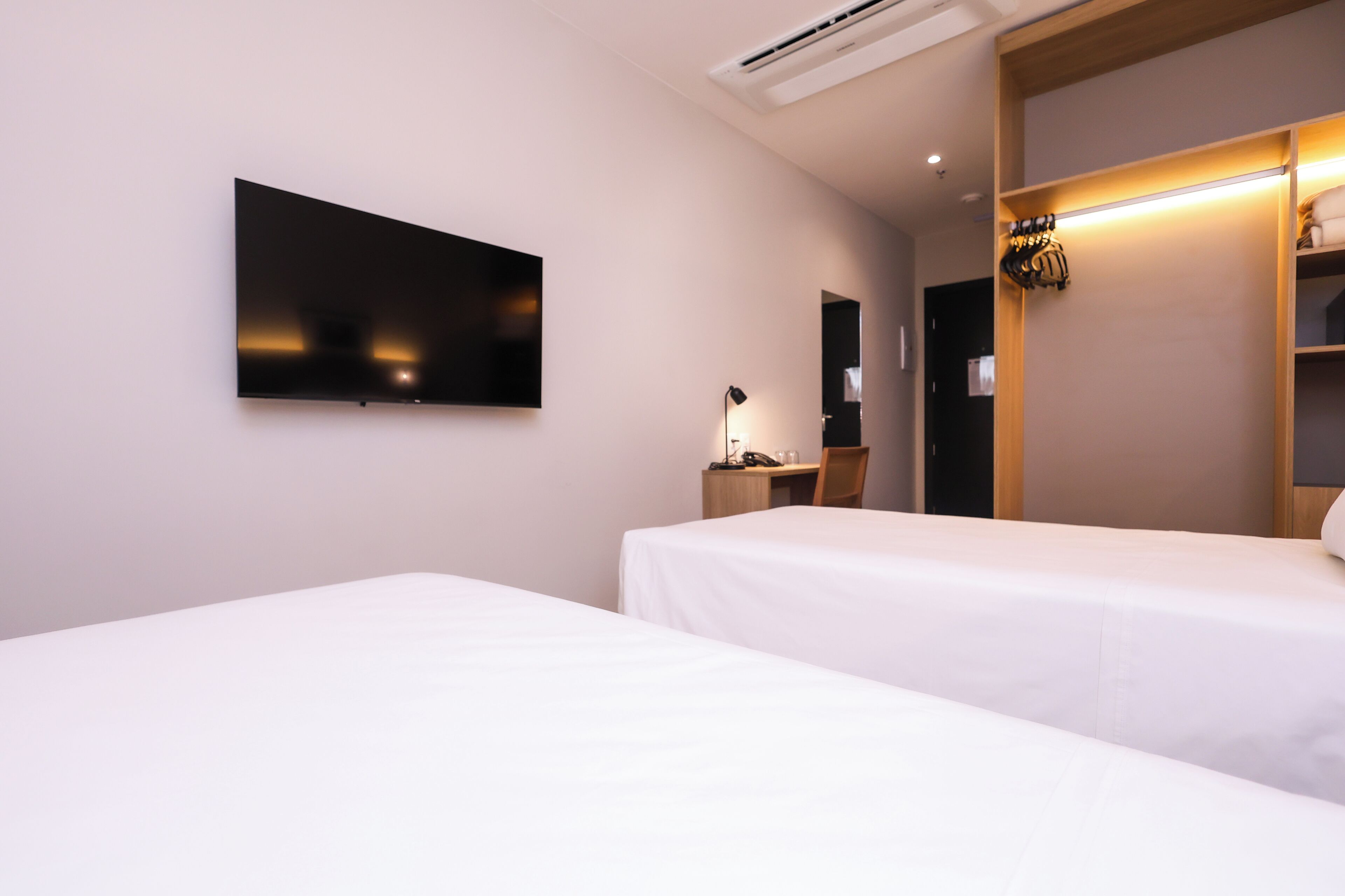 A hotel room with two beds, a flat-screen TV mounted on the wall, a desk with a lamp, and a wardrobe with a mirror.