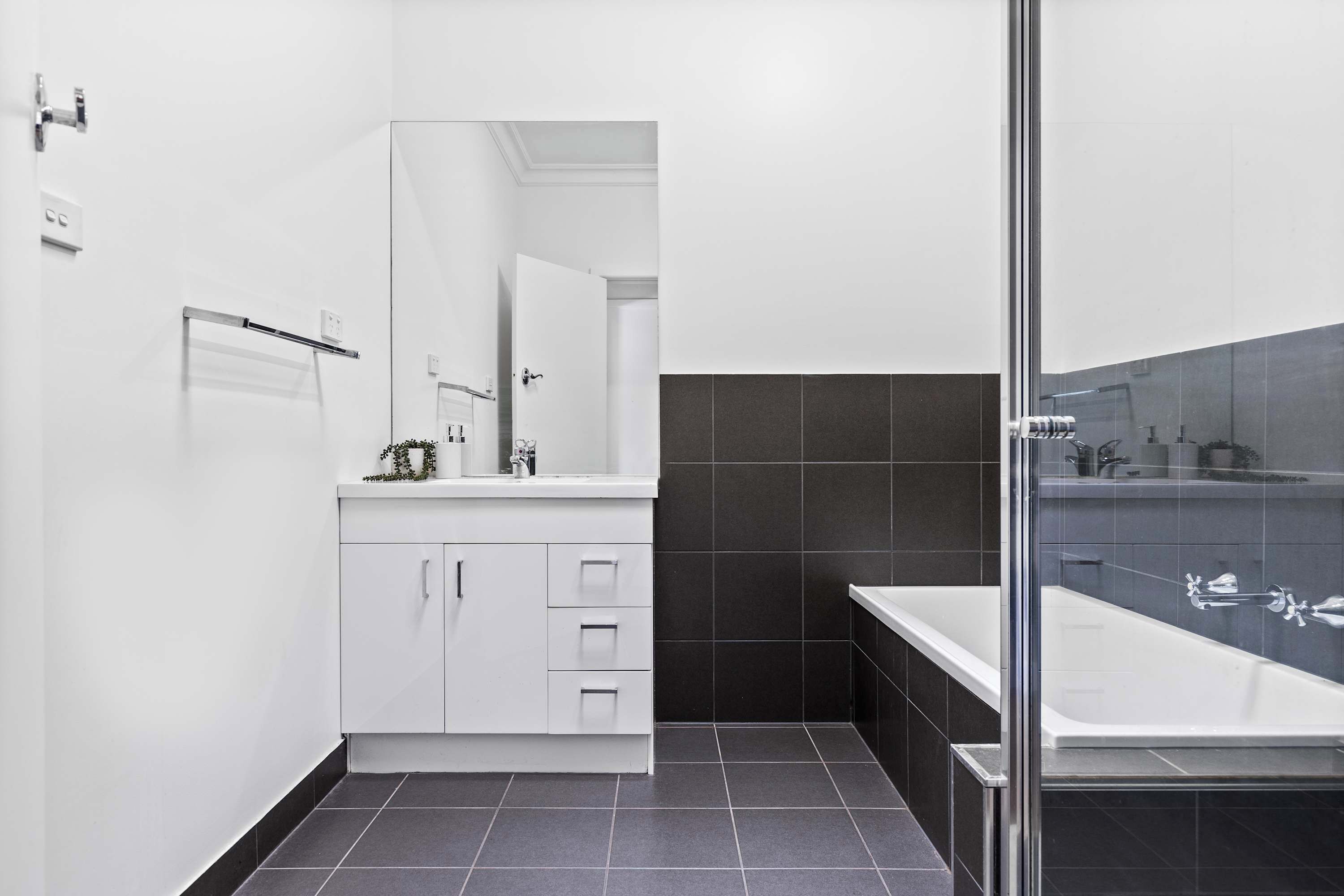 Une salle de bain moderne avec un meuble-lavabo blanc, un grand miroir, un mur recouvert de carreaux foncés et une douche séparée par une paroi de verre.