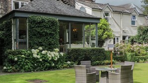 Een oranjerie met glazen dak, omringd door weelderige begroeiing en witte bloemen.