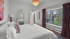 A spacious bedroom with a large bed, a chandelier, a view of the outdoors, and a window with curtains. - Eventide: Renovated 5 Bedroom, 3 Baths on Aquidneck Island (Portsmouth)