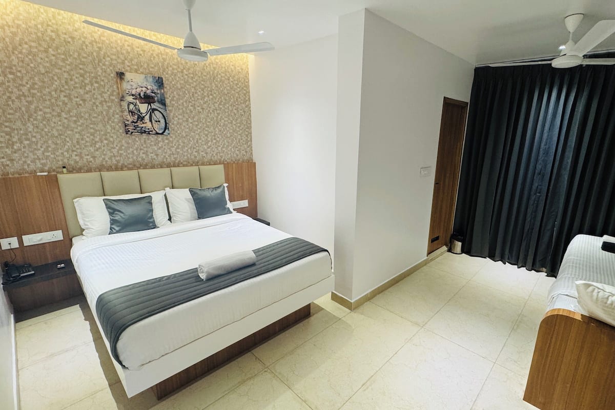 A hotel room with a bed, a bedside table, a ceiling fan, a painting on the wall, and a window with curtains.