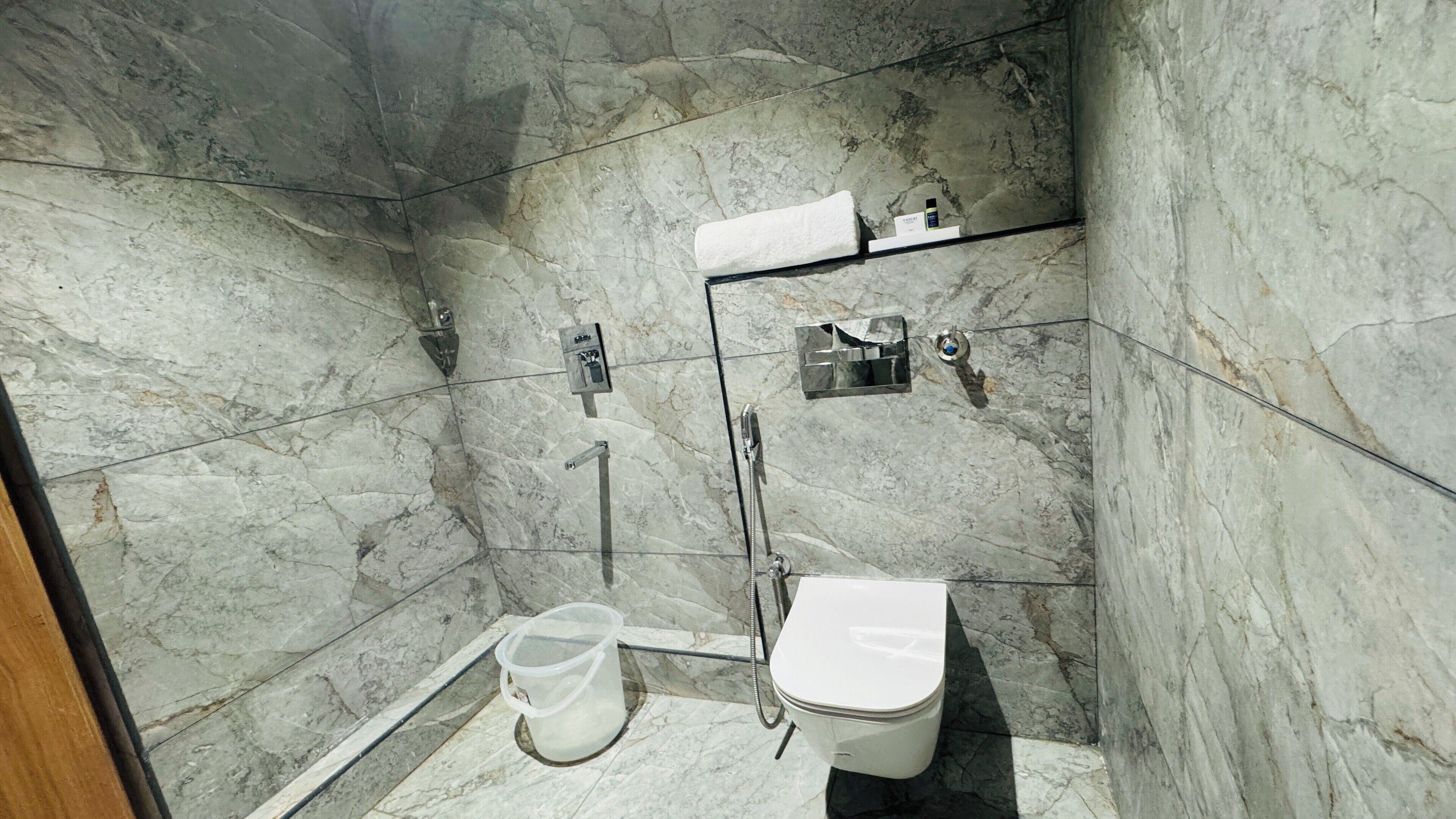 A modern bathroom with a marble floor, a toilet, a shower, and a small shelf with toiletries.