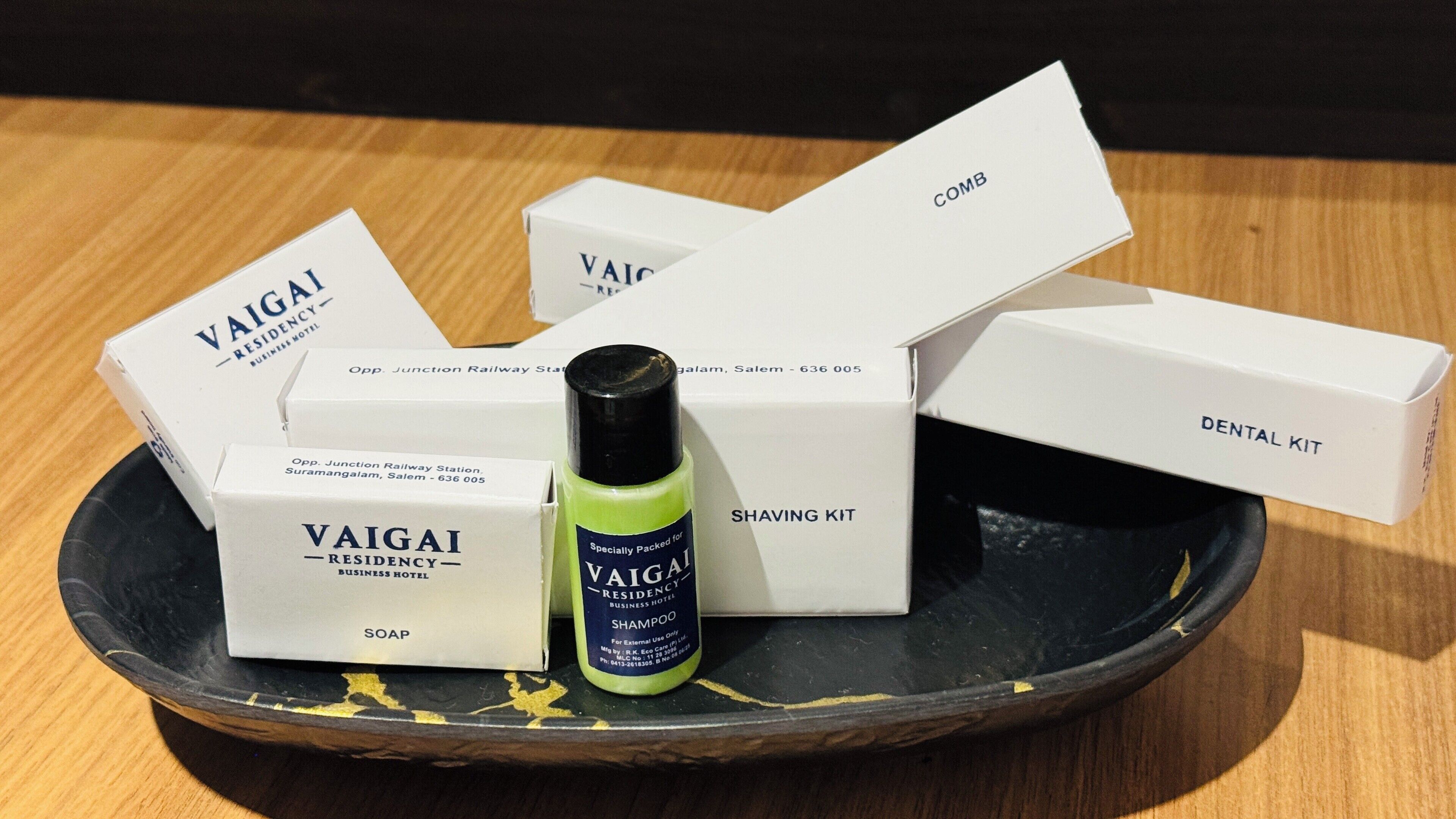 Una bandeja negra con varias cajas blancas etiquetadas 'VAIGAI RESIDENCY BUSINESS HOTEL' y una botella verde de champú.