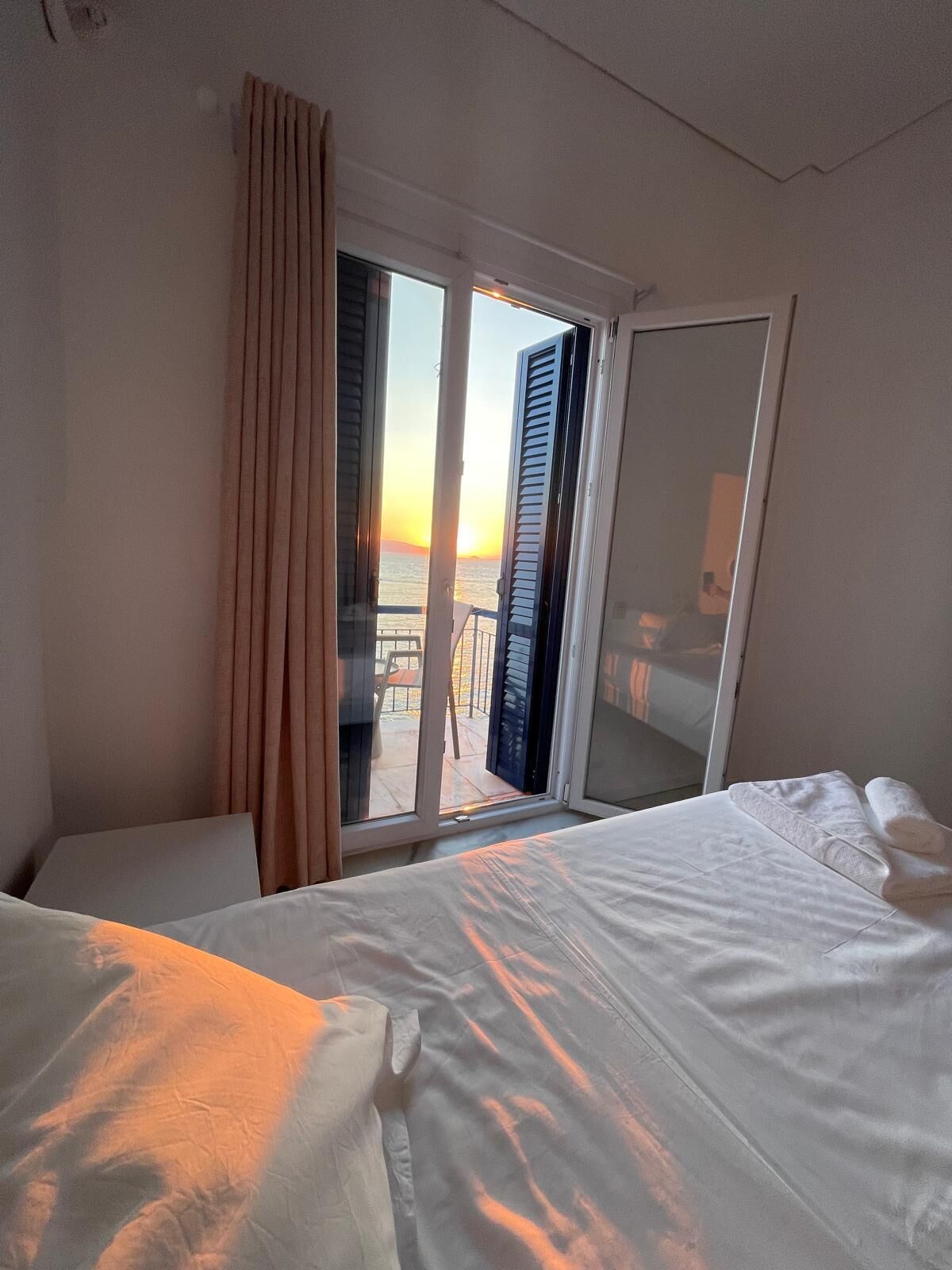 Une chambre d’hôtel avec un lit, une chaise, une fenêtre avec des rideaux et une vue sur la mer au coucher du soleil.