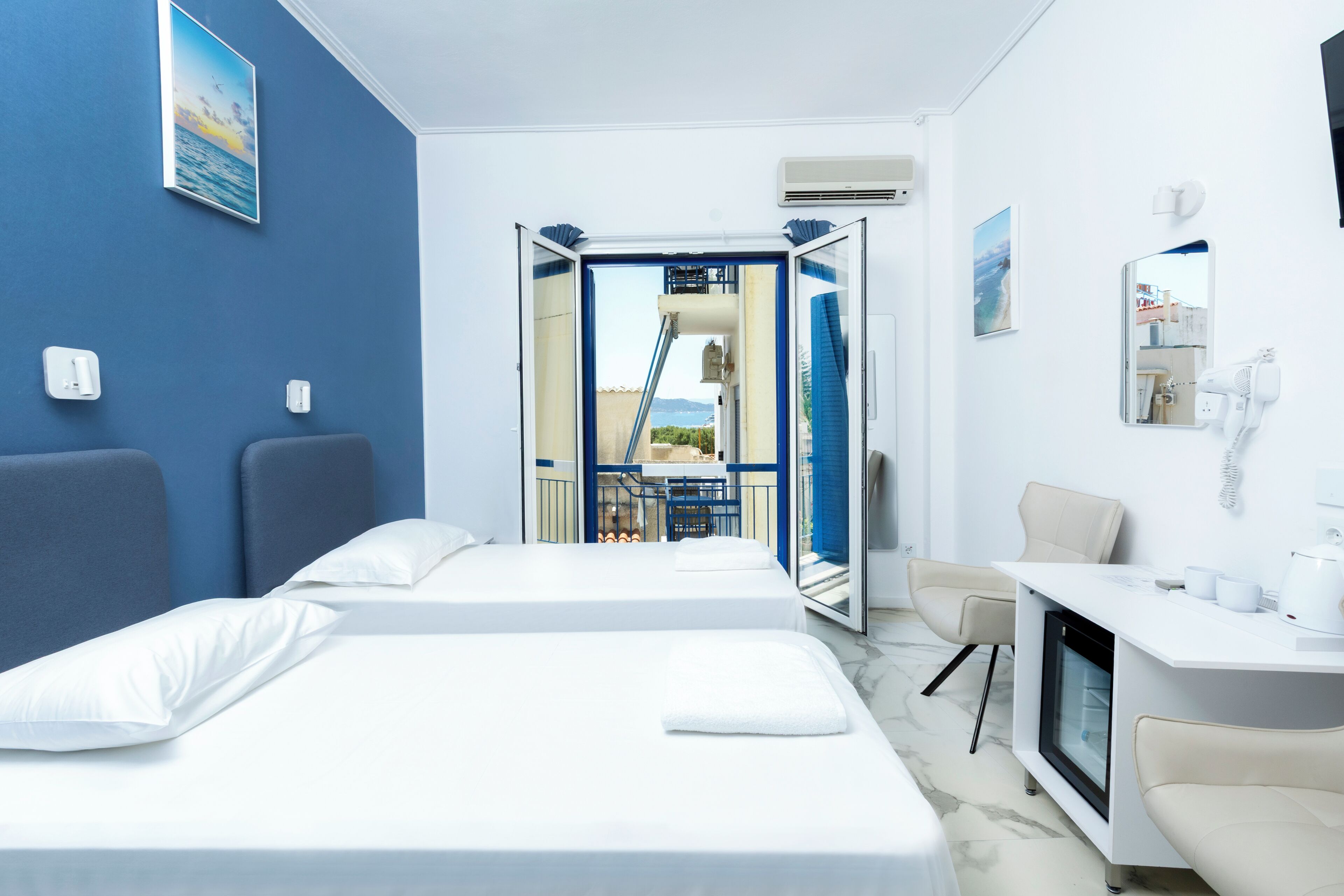 A hotel room with two beds, a desk, a chair, a small table, a mirror, and a view of the outside.