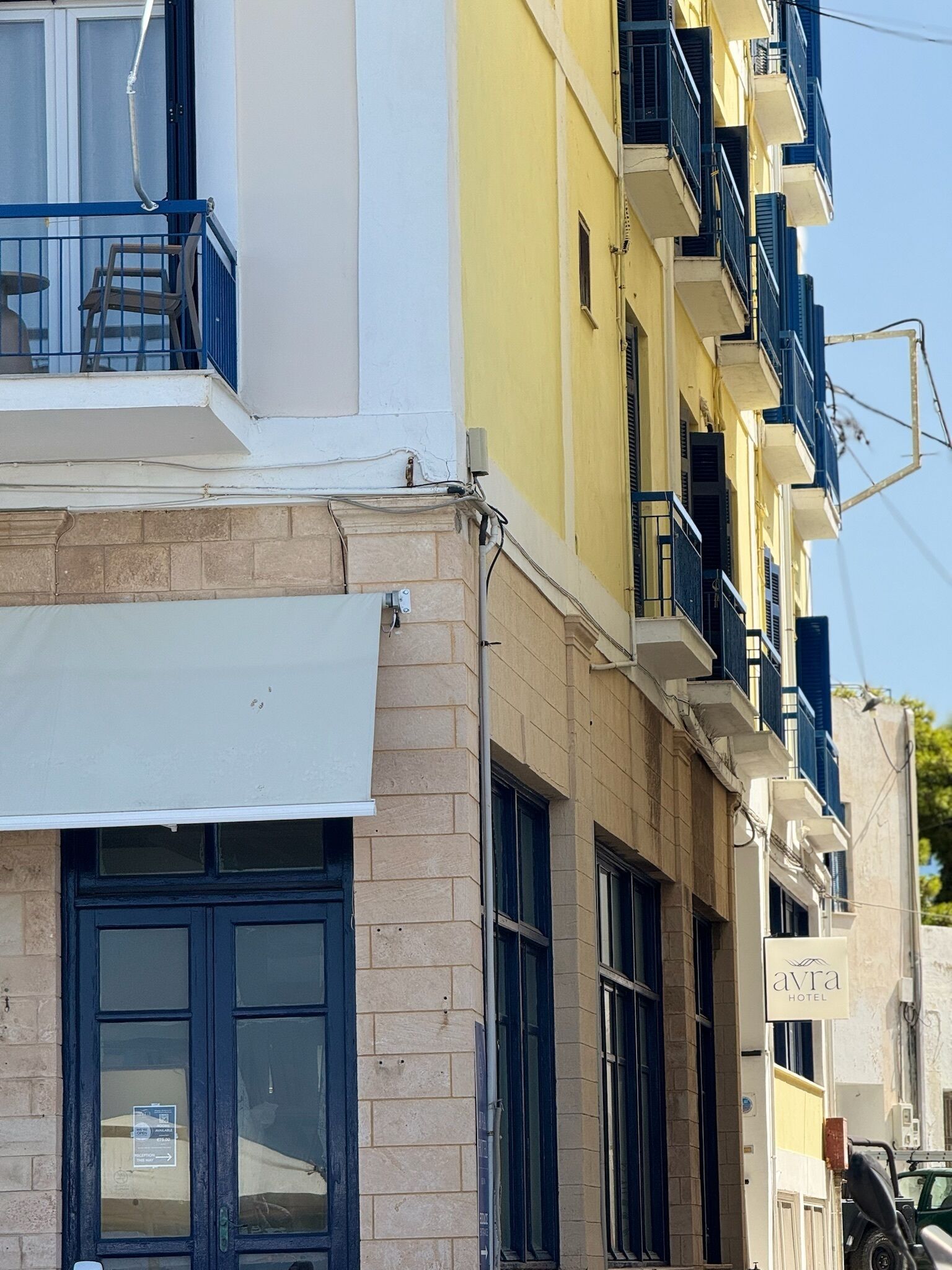 A hotel facade with a white awning, a sign reading "Avra Hotel," and a vehicle parked outside.