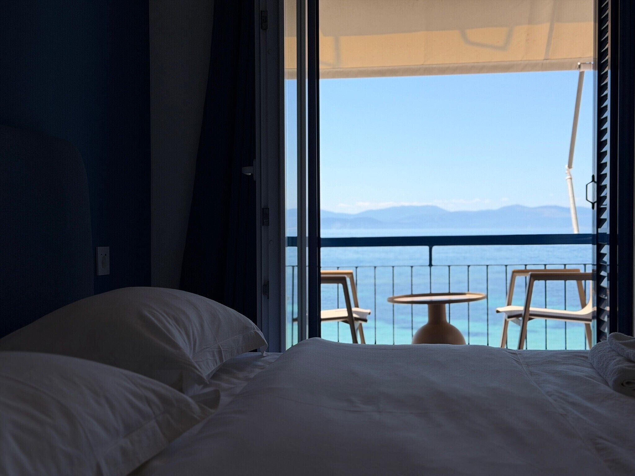 Une chambre d’hôtel avec un lit, une fenêtre donnant sur la mer et un balcon avec des chaises.