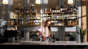 Zona de bar con estantes llenos de botellas de licor, una barra y una persona preparando una bebida.