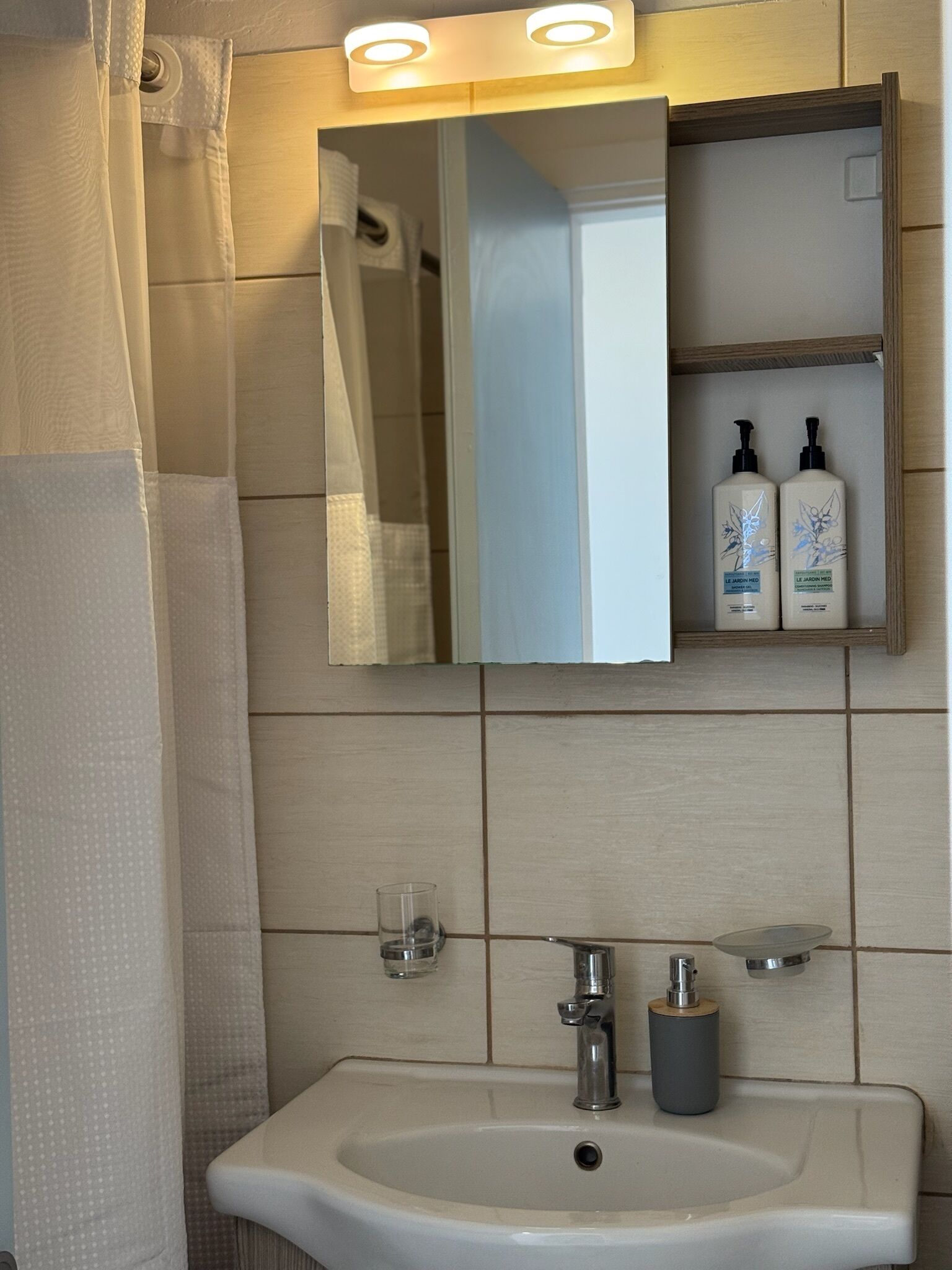 Une salle de bain avec un lavabo blanc, un miroir et une étagère sur laquelle sont posées deux bouteilles de Le Jardin Med.