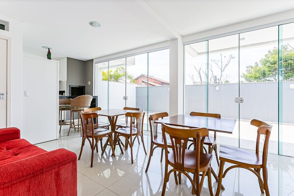 A modern dining area with wooden tables and chairs, a red sofa, and large windows offering a view of the outside. - New Studio 900 m From Lagoa da Conceicao - Ecm204 (Florianópolis)