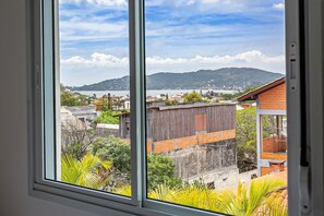 A view from a window overlooking a coastal area with buildings, greenery, and a distant mountain range. - New Studio 900 m From Lagoa da Conceicao - Ecm204 (Florianópolis)