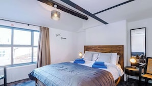 A bedroom with a large bed, a wooden headboard, a bedside table with a lamp, a chair, and a window with curtains.